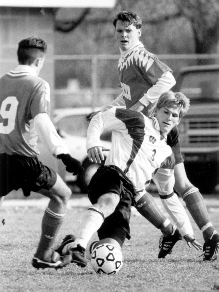 Black-and-white photograph of a soccer player sliding to kick the ball, with two opposing players close by.
