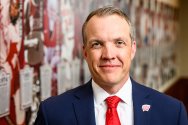 A man in a blue suit, red patterned tie and Motion W lapel pin, smiles in a head-and-shoulders portrait, standing in front of a blurred wall display featuring Badger football imagery.