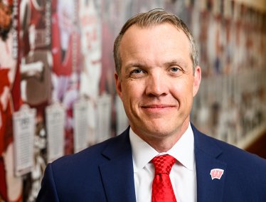 A man in a blue suit, red patterned tie and Motion W lapel pin, smiles in a head-and-shoulders portrait, standing in front of a blurred wall display featuring Badger football imagery.