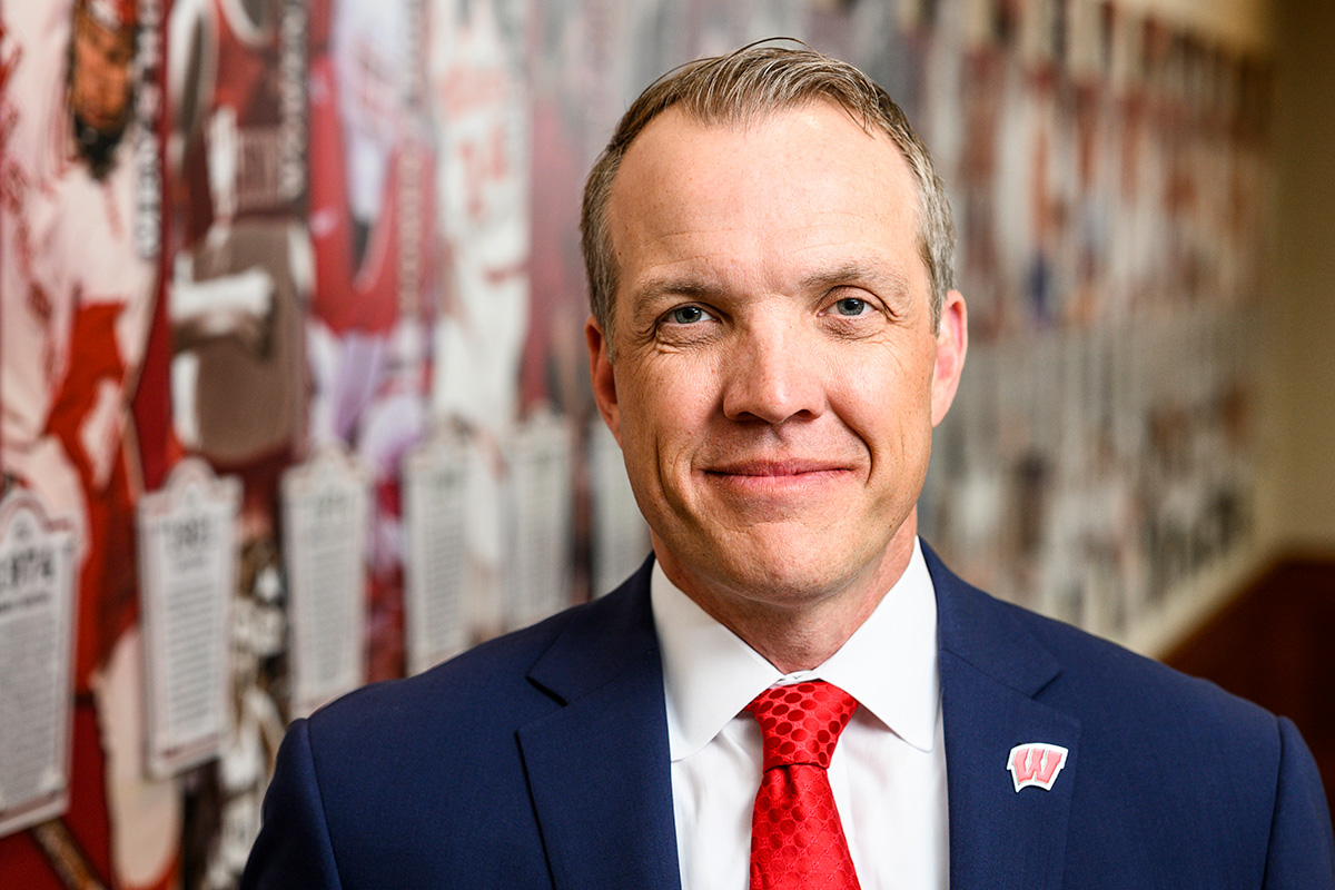 A man in a blue suit, red patterned tie and Motion W lapel pin, smiles in a head-and-shoulders portrait, standing in front of a blurred wall display featuring Badger football imagery.