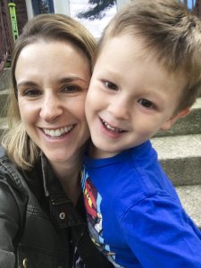 A happy young boy in a blue t-shirt and shorts hugs his mom and looks at the camera.
