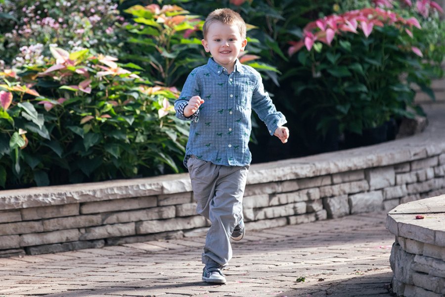 A smiling boy wearing a blue button-down shirt runs down a walkway with flowering shrubs behind him.