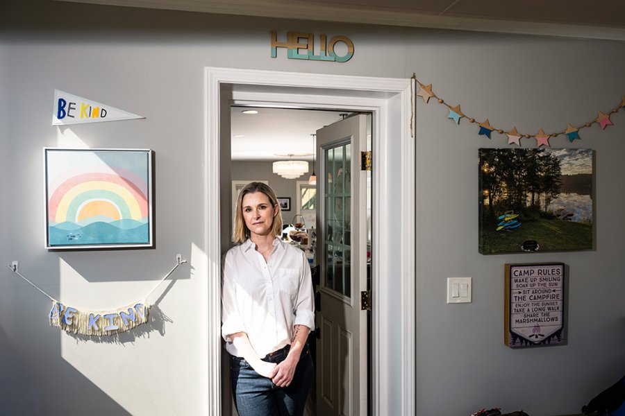 A woman standing in a doorway inside a home decorated with colorful wall art and signs, including a 'Hello' sign above the door, a 'Be Kind' banner, and framed artwork on either side.