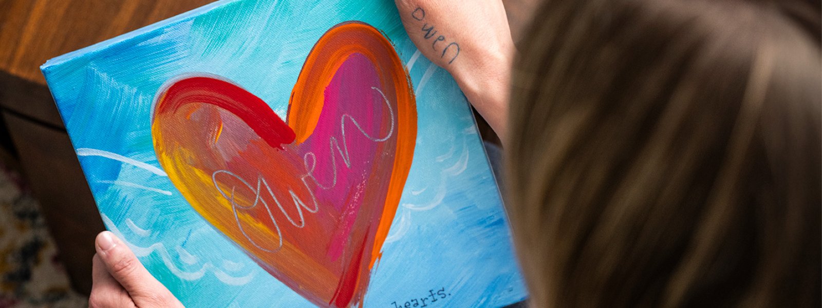 Person holding a colorful painted canvas featuring a large heart design with the word 'Owen' and the phrase 'forever in our hearts'.