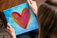 Person holding a colorful painted canvas featuring a large heart design with the word 'Owen' and the phrase 'forever in our hearts'.