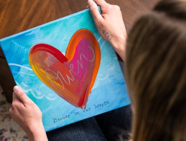 Person holding a colorful painted canvas featuring a large heart design with the word 'Owen' and the phrase 'forever in our hearts'.