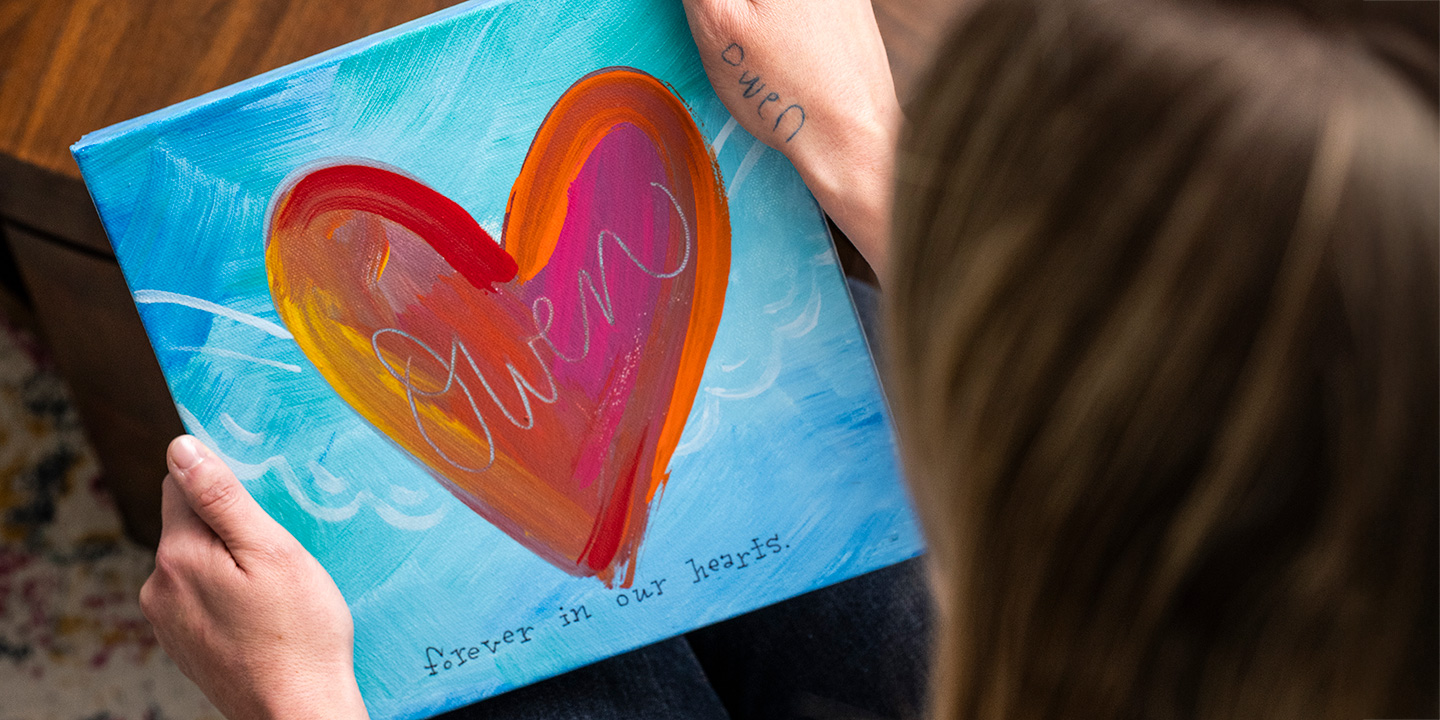 Person holding a colorful painted canvas featuring a large heart design with the word 'Owen' and the phrase 'forever in our hearts'.
