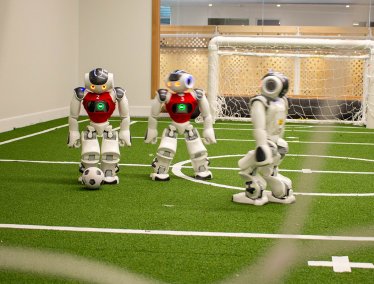 Three small humanoid robots play soccer on a miniature green turf field with white markings and a small goal in the background.