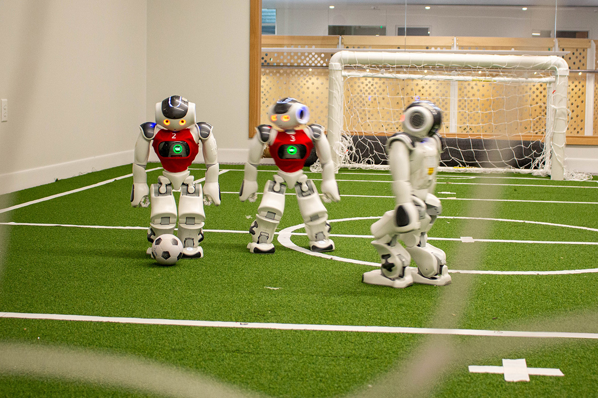 Three small humanoid robots play soccer on a miniature green turf field with white markings and a small goal in the background.