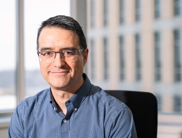 A professional headshot of a smiling man with short dark hair and glasses, wearing a blue patterned button-down shirt, sitting indoors with a blurred building exterior visible through a window in the background.