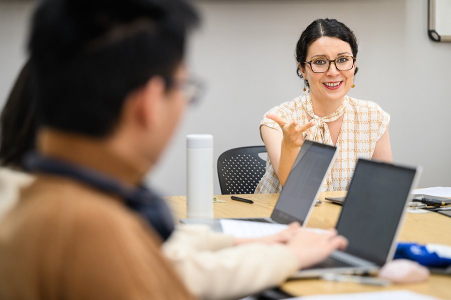 Toma, seated at a table, gestures while speaking during a classroom discussion as others work on laptops nearby.