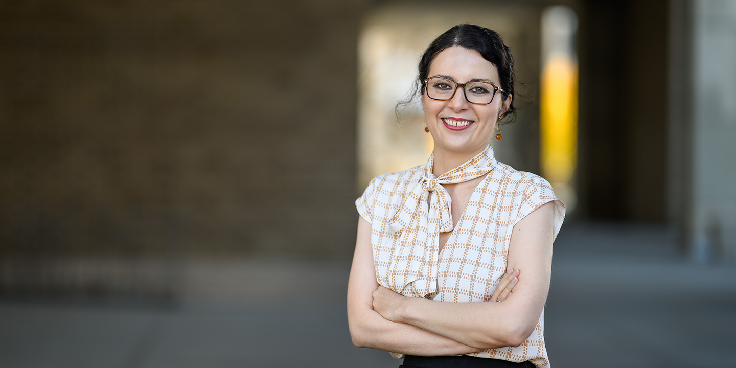 A woman wearing glasses is depicted, with arms crossed in an outdoor walkway, with blurred architectural columns and soft lighting in the background.
