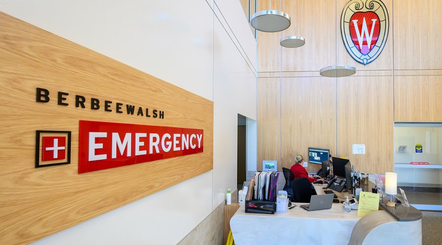 A bright, modern reception area for Berbee Walsh Emergency featuring a light wood-paneled wall, a curved white desk where a staff member works, and a large stained-glass University of Wisconsin 'W' crest.