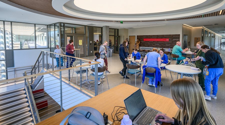 A high-angle view of a bustling student lounge and cafe area with people sitting at round tables, a woman working on a laptop in the foreground, and a large staircase leading to a lower level.