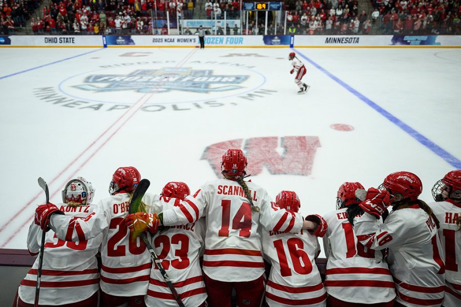 Group of hockey players in white and red uniforms lean together along the boards watching their teammate on the ice during the NCAA Women’s Frozen Four championship game.