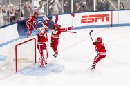 Ice hockey players in red uniforms celebrate near the goal crease on the ice while spectators cheer behind the rink boards.