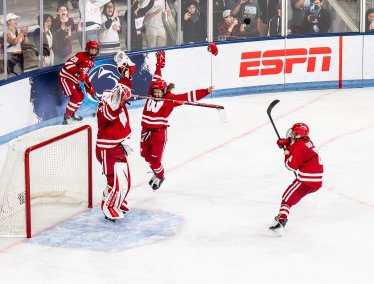 Ice hockey players in red uniforms celebrate near the goal crease on the ice while spectators cheer behind the rink boards.