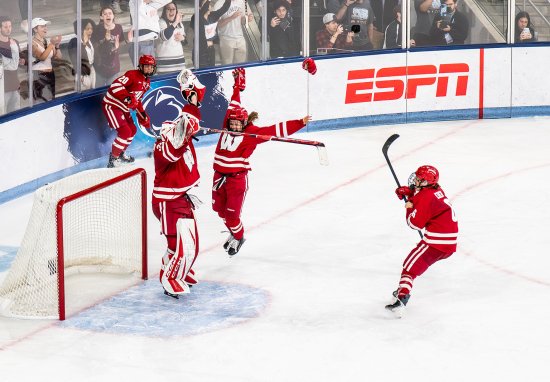 Ice hockey players in red uniforms celebrate near the goal crease on the ice while spectators cheer behind the rink boards.