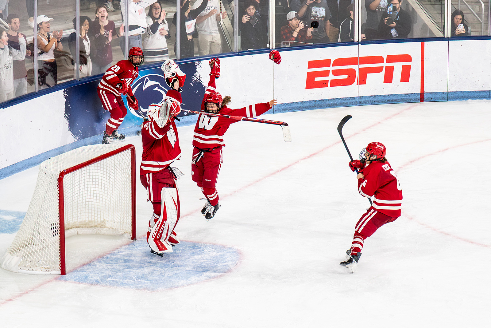 Ice hockey players in red uniforms celebrate near the goal crease on the ice while spectators cheer behind the rink boards.
