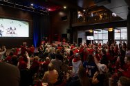 A large crowd wearing red gathers inside an event space to watch an ice hockey game on a large screen, cheering, waving flags, and raising hands as the action unfolds.