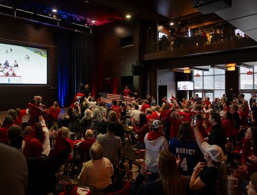 A large crowd wearing red gathers inside an event space to watch an ice hockey game on a large screen, cheering, waving flags, and raising hands as the action unfolds.