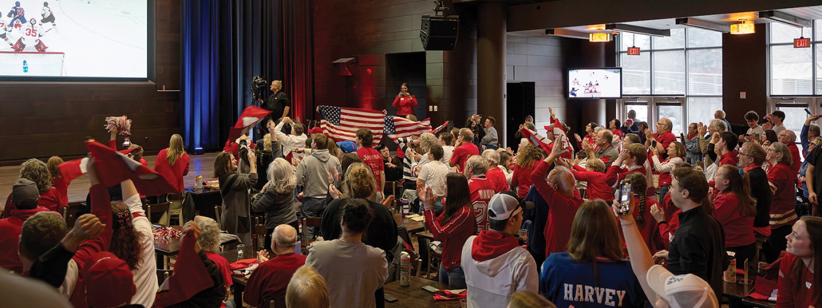 A wide view shows a packed indoor venue of spectators in red apparel waving flags and raising hands while watching the UW Badgers' ice hockey game on multiple screens.