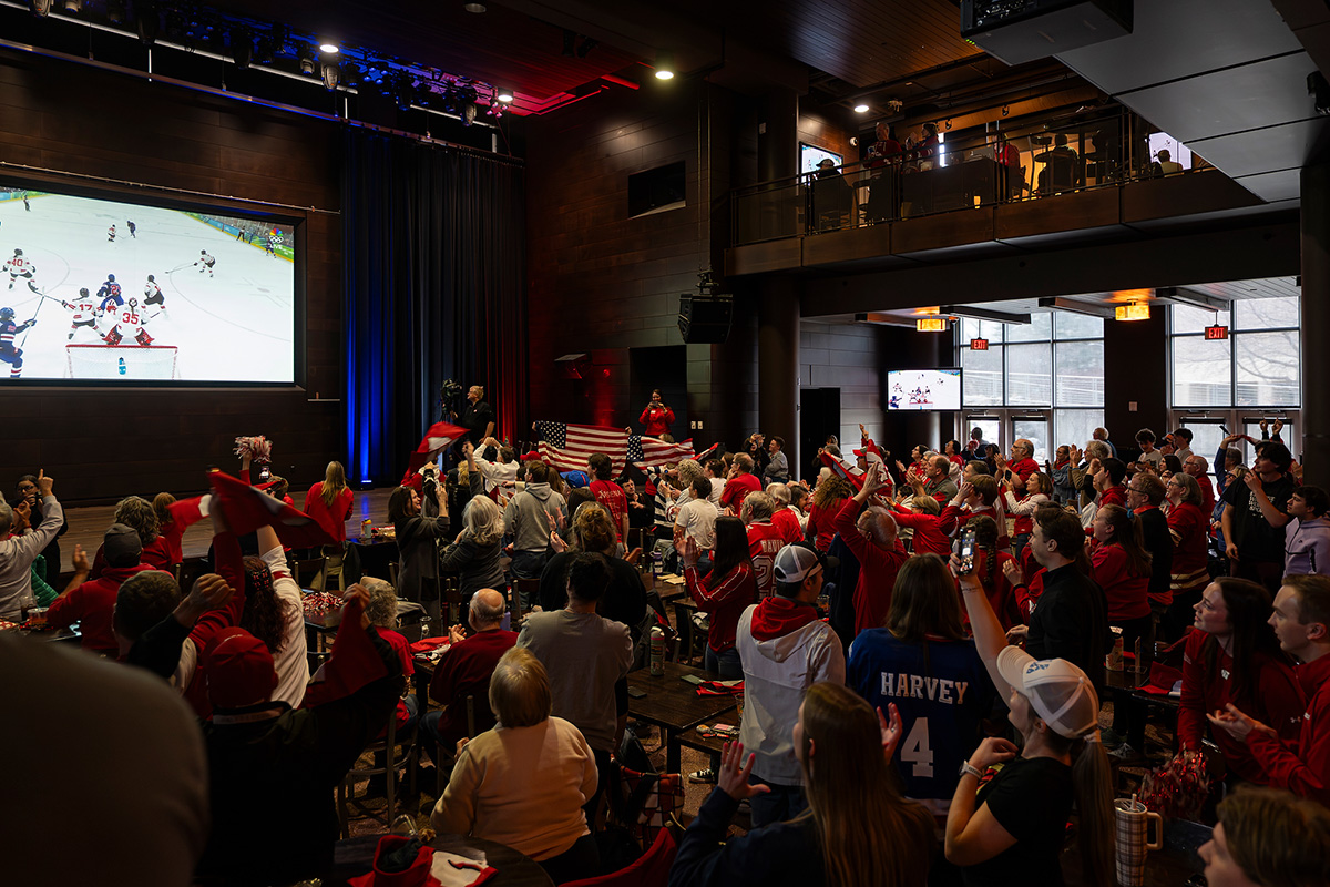 A large crowd wearing red gathers inside an event space to watch an ice hockey game on a large screen, cheering, waving flags, and raising hands as the action unfolds.
