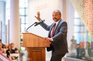 Eric Wilcots, wearing a gray suit and red tie, speaks from a wooden podium with his right hand raised during a presentation in a brightly lit campus building.