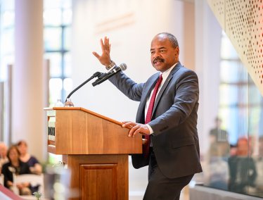 Eric Wilcots, wearing a gray suit and red tie, speaks from a wooden podium with his right hand raised during a presentation in a brightly lit campus building.