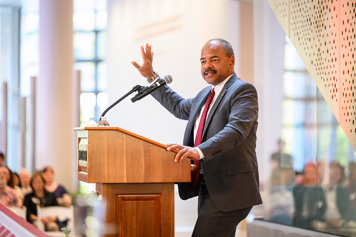 Eric Wilcots, wearing a gray suit and red tie, speaks from a wooden podium with his right hand raised during a presentation in a brightly lit campus building.