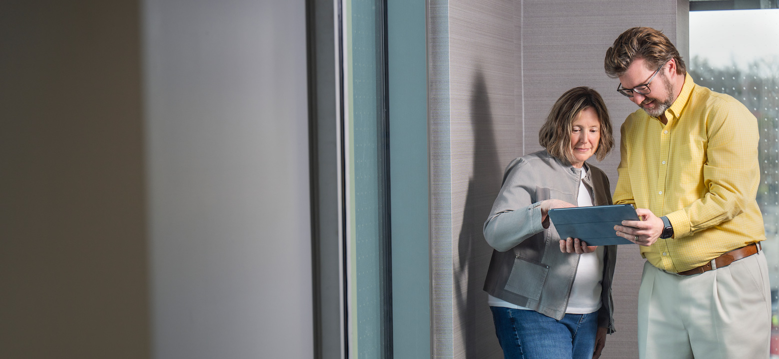 Two people stand near a window and look at a tablet together, with one person pointing at the screen in a softly lit interior space.