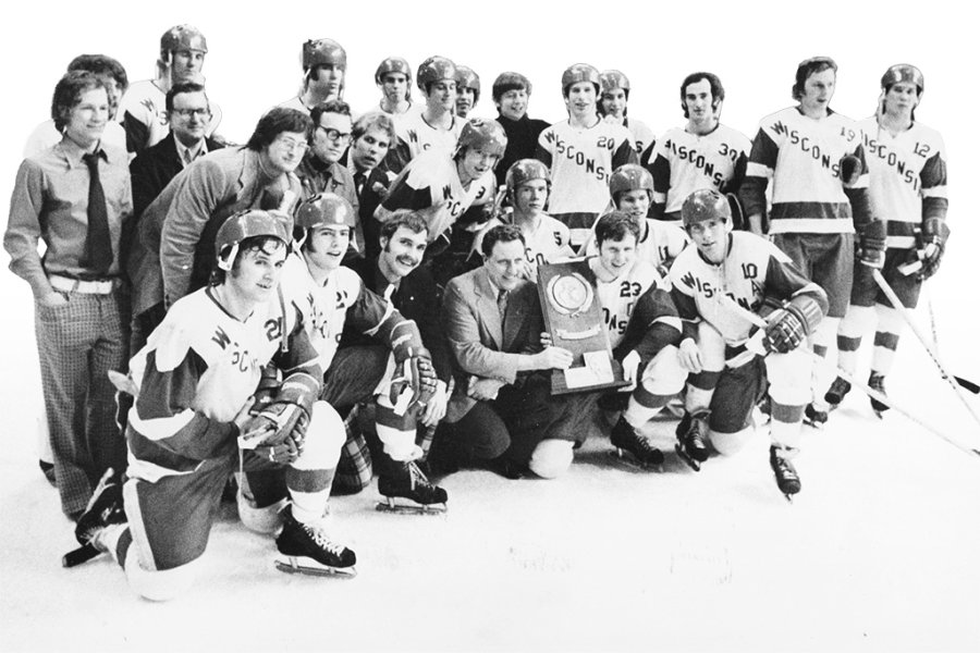 Black-and-white photo of a hockey team in Wisconsin uniforms gathered on the ice with coaches and staff, posing together while one person holds a championship trophy at the center.