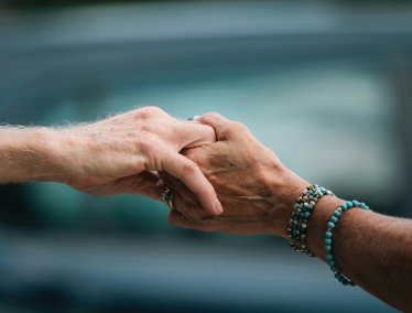 A close-up shot of two elderly hands gently holding each other, with one person wearing beaded bracelets, set against a softly blurred greenish-blue background.