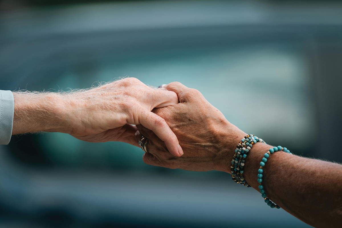 A close-up shot of two elderly hands gently holding each other, with one person wearing beaded bracelets, set against a softly blurred greenish-blue background.