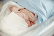 A newborn baby wearing a white beanie sleeps soundly while swaddled in a light blue blanket inside a hospital bassinet.