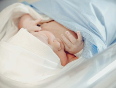 A newborn baby wearing a white beanie sleeps soundly while swaddled in a light blue blanket inside a hospital bassinet.