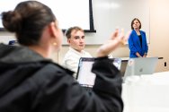 A professor stands at the front of a lecture hall while one student gestures while talking, and another student listens.