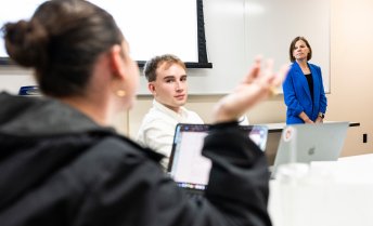 A professor stands at the front of a lecture hall while one student gestures while talking, and another student listens.
