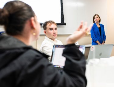 A professor stands at the front of a lecture hall while one student gestures while talking, and another student listens.
