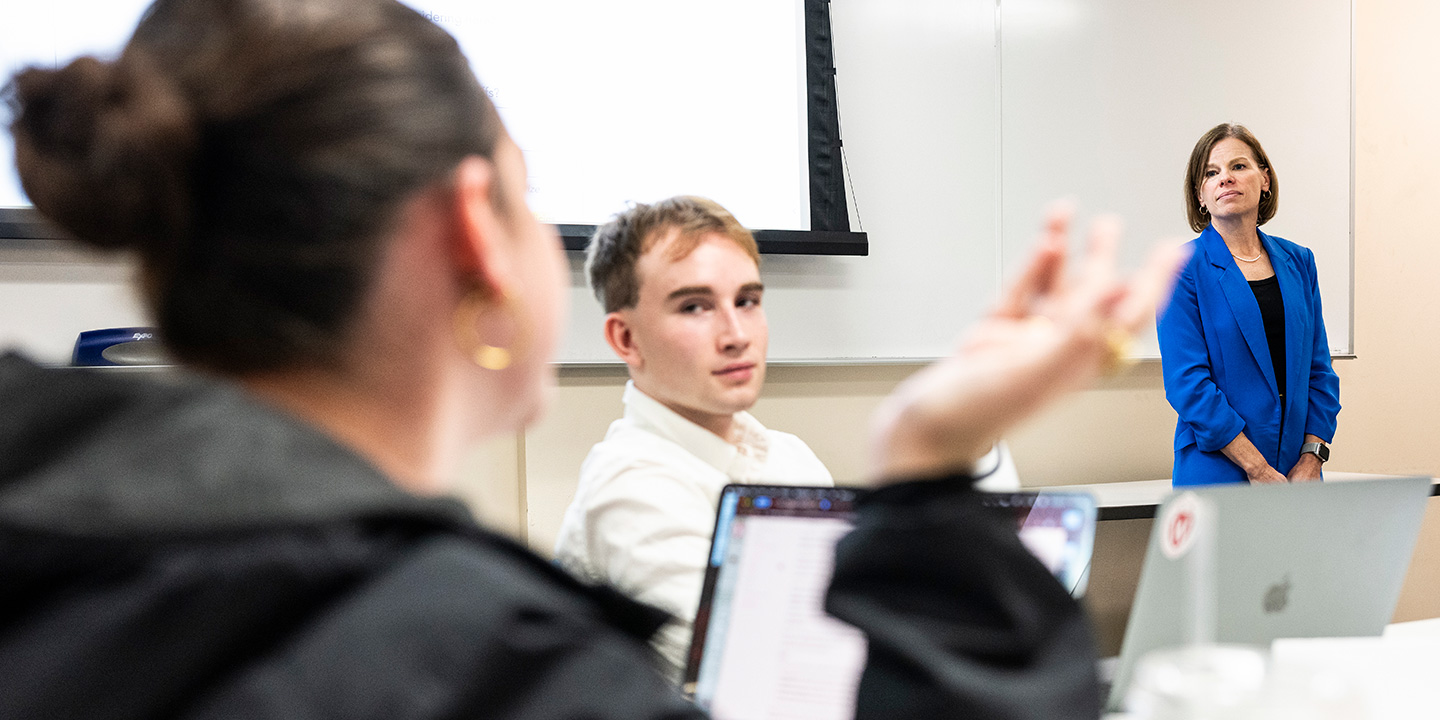 A professor stands at the front of a lecture hall while one student gestures while talking, and another student listens.