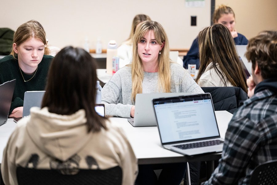 Small group of students sit around a table in a classroom working on laptops while one student speaks as others listen and take notes.