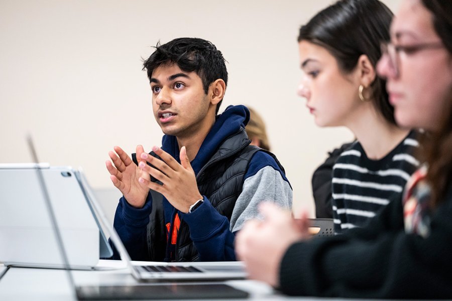 A person at a table speaks while gesturing with both hands during a classroom discussion, with other students seated nearby use laptops and listen.