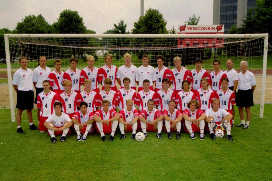 A slightly yellowed photo of a team of soccer players and coaches in matching uniforms posing in front of a goal on a grass field, with a scoreboard displaying 'Wisconsin' in the background.