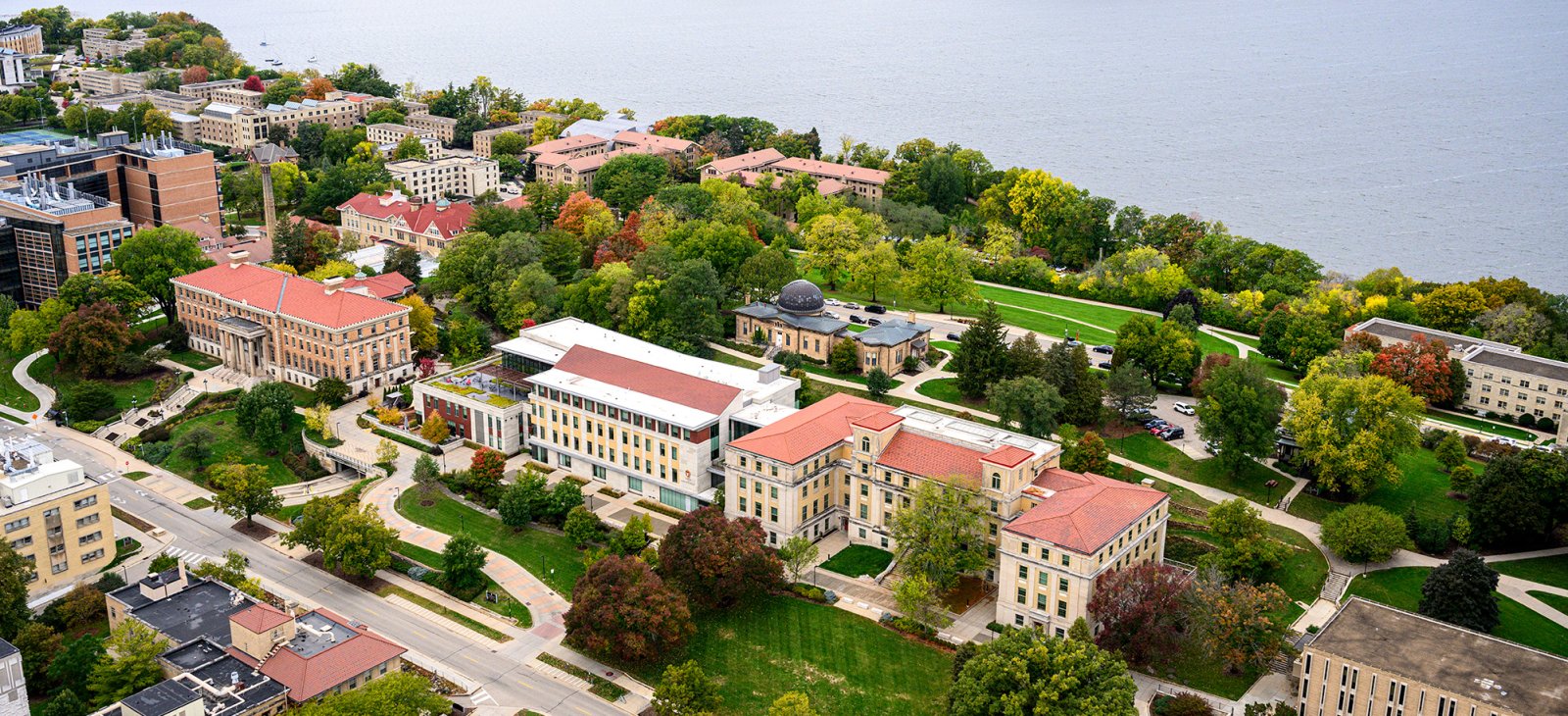 An aerial view of university campus buildings with red-tiled roofs and autumn foliage along the shoreline of a lake.