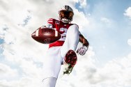 A low-angle shot of a Wisconsin Badgers football player high-stepping toward the camera and extending a football against a bright, cloudy blue sky.