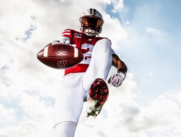 A low-angle shot of a Wisconsin Badgers football player high-stepping toward the camera and extending a football against a bright, cloudy blue sky.