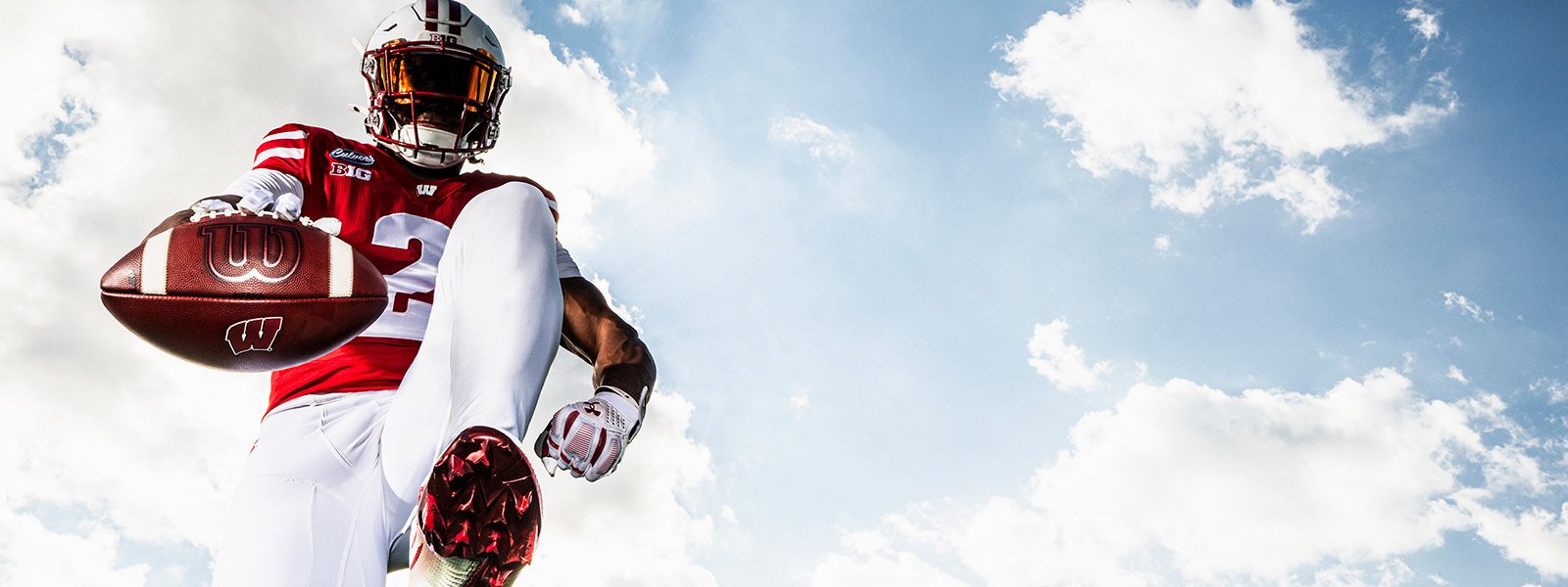 A low-angle shot of a Wisconsin Badgers football player high-stepping toward the camera and extending a football against a bright, cloudy blue sky.