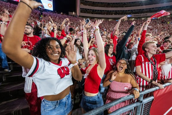 Enthusiastic fans wearing red and white Wisconsin apparel cheer with raised arms in the stadium stands.