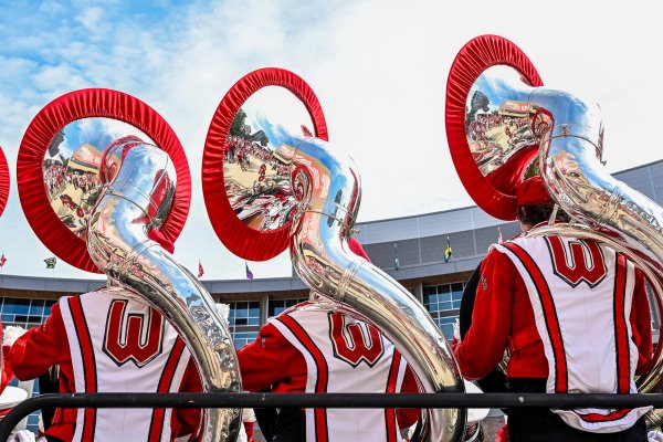 Three marching band members in red and white uniforms stand facing away from the camera while holding silver sousaphones trimmed with red covers.