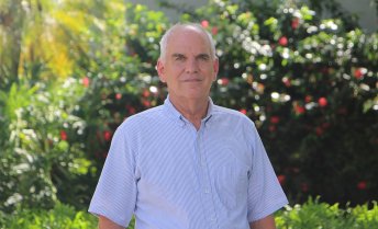 Portrait of Bruce McCord wearing a short-sleeved blue pinstriped shirt, standing outdoors in front of lush green foliage.